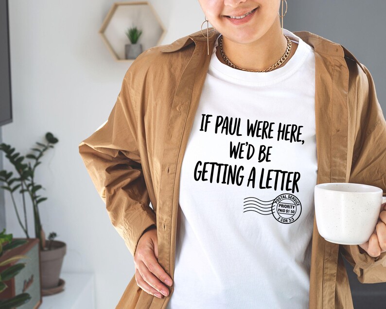Puede incluir: Camiseta blanca con el texto "IF PAUL WERE HERE, WE'D BE GETTING A LETTER" en negro. La camiseta tambi&eacute;n presenta un gr&aacute;fico de sello postal. La persona lleva una chaqueta marr&oacute;n y sostiene una taza blanca.