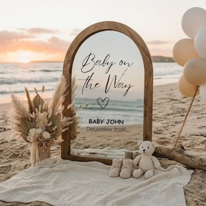 May include: A wooden-framed mirror with the text "Baby on the Way" and "Baby John December 2026" on a beach. A teddy bear, baby booties, and a bouquet of dried flowers sit on a blanket. Balloons are tied to a piece of driftwood.
