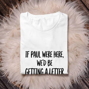Puede incluir: Una camiseta blanca con el texto negro "IF PAUL WERE HERE, WE'D BE GETTING A LETTER". La camiseta est&aacute; doblada y descansa sobre una superficie blanca y esponjosa, con un fondo de madera.