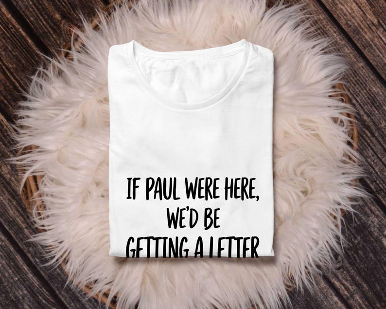 Puede incluir: Una camiseta blanca con el texto negro "IF PAUL WERE HERE, WE'D BE GETTING A LETTER". La camiseta est&aacute; doblada y descansa sobre una superficie blanca y esponjosa, con un fondo de madera.