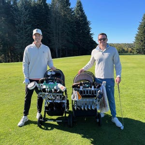 May include: Two golf carts filled with golf clubs on a green golf course. The carts are black with brown accents. Two people are standing next to the carts. The sky is blue and there are tall green trees in the background.