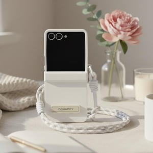 May include: A cream-coloured phone case with a braided strap, bearing the text "QGHAPPY" on a small tag. The phone case is open, displaying a black screen. A pink flower and a candle are in the background.
