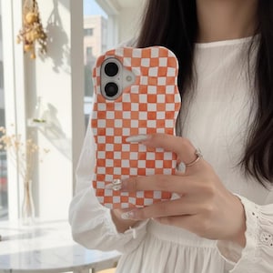 Könnte beinhalten: Eine Handyhülle mit gewelltem Rand, die ein kariertes Muster in Orange und Weiß aufweist. Die Hülle hat Aussparungen für die Kamera und die Tasten des Telefons. Die Handyhülle wird vor einem weißen Kleid gehalten.