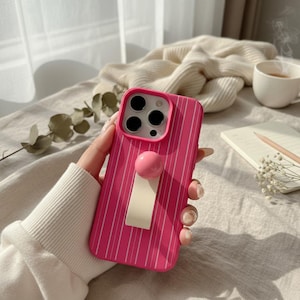 May include: A bright pink phone case with vertical white stripes. The case has a pink ball and a cream-colored rectangular grip. The phone case is held by a person wearing a white sweater.