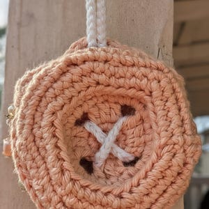 May include: A peach-colored, crocheted button-shaped item with white thread detailing. The button has four holes with white thread crisscrossing through them. A white braided cord is attached for hanging.
