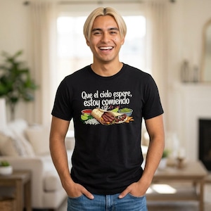 May include: A black short-sleeved t-shirt featuring a graphic of a burrito and taco with the text "Que el cielo espere, estoy comiendo" printed in white. The design includes small bowls of salsa, guacamole, and lime wedges surrounding the food.