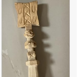 May include: A carved wooden Oshe Sango dance wand. The wand features intricate carvings, including a rectangular top with leaf-like designs, a figure with arms crossed, and geometric shapes. The wand is made of light-coloured wood.