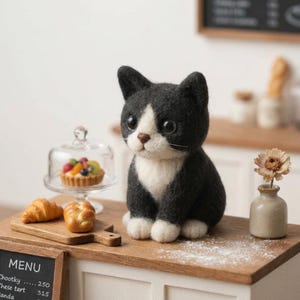 May include: A needle-felted black and white cat figurine with large dark eyes, sitting on a wooden counter next to a miniature glass cloche containing a fruit tart, a small wooden board with two pastries, and a tiny vase with a dried flower.