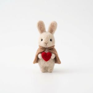 May include: A handmade, beige felt bunny figurine with a brown cape, holding a red heart. The bunny has long ears and a sweet expression. The background is plain white.