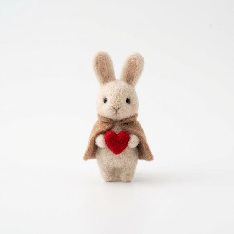 May include: A handmade, beige felt bunny figurine with a brown cape, holding a red heart. The bunny has long ears and a sweet expression. The background is plain white.