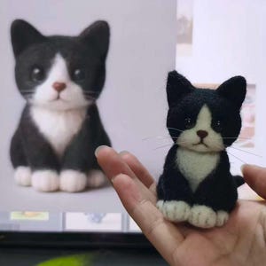 May include: A small needle-felted cat figurine with black and white fur, featuring a white chest, white paws, and a black face with a white blaze. The cat has dark eyes, a brown nose, and thin, long whiskers extending from its muzzle.
