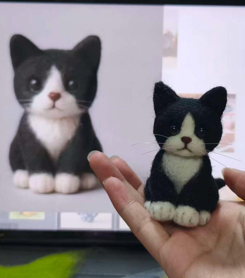 May include: A small needle-felted cat figurine with black and white fur, featuring a white chest, white paws, and a black face with a white blaze. The cat has dark eyes, a brown nose, and thin, long whiskers extending from its muzzle.