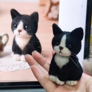 May include: A small, needle-felted black and white cat figurine with a white chest, white paws, and a white blaze on its face. The cat has dark, round eyes, a small brown nose, and long, thin white whiskers, appearing as a miniature sculpture.