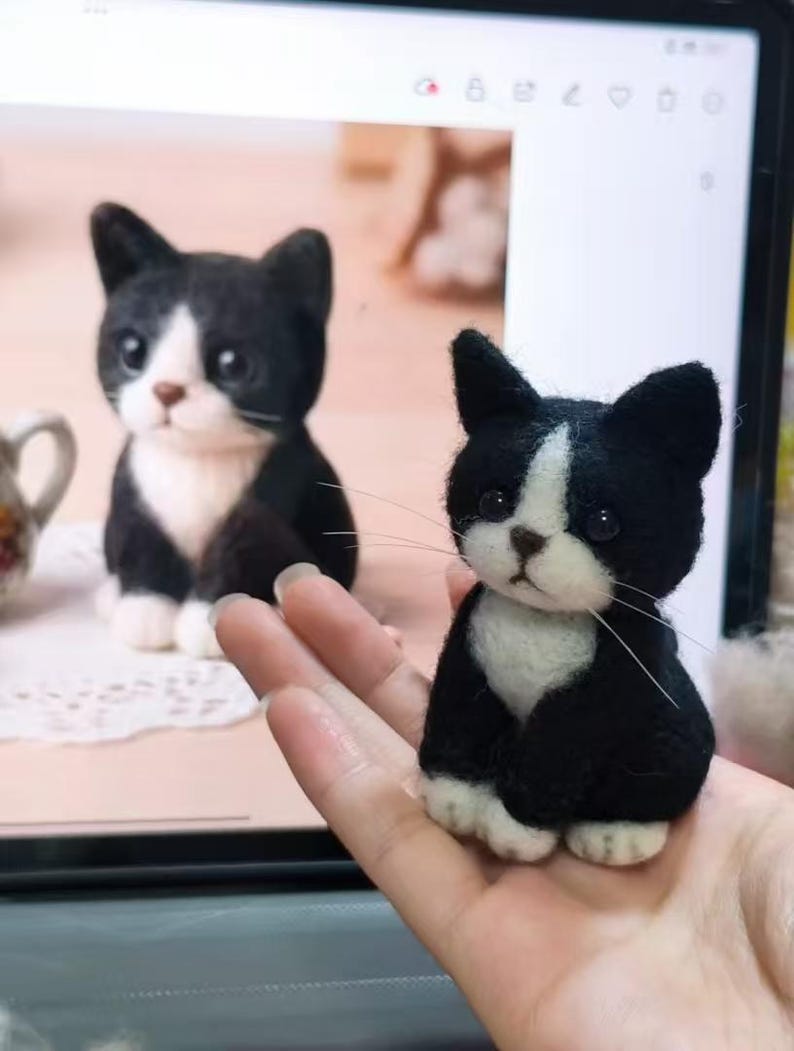 May include: A small, needle-felted black and white cat figurine with a white chest, white paws, and a white blaze on its face. The cat has dark, round eyes, a small brown nose, and long, thin white whiskers, appearing as a miniature sculpture.