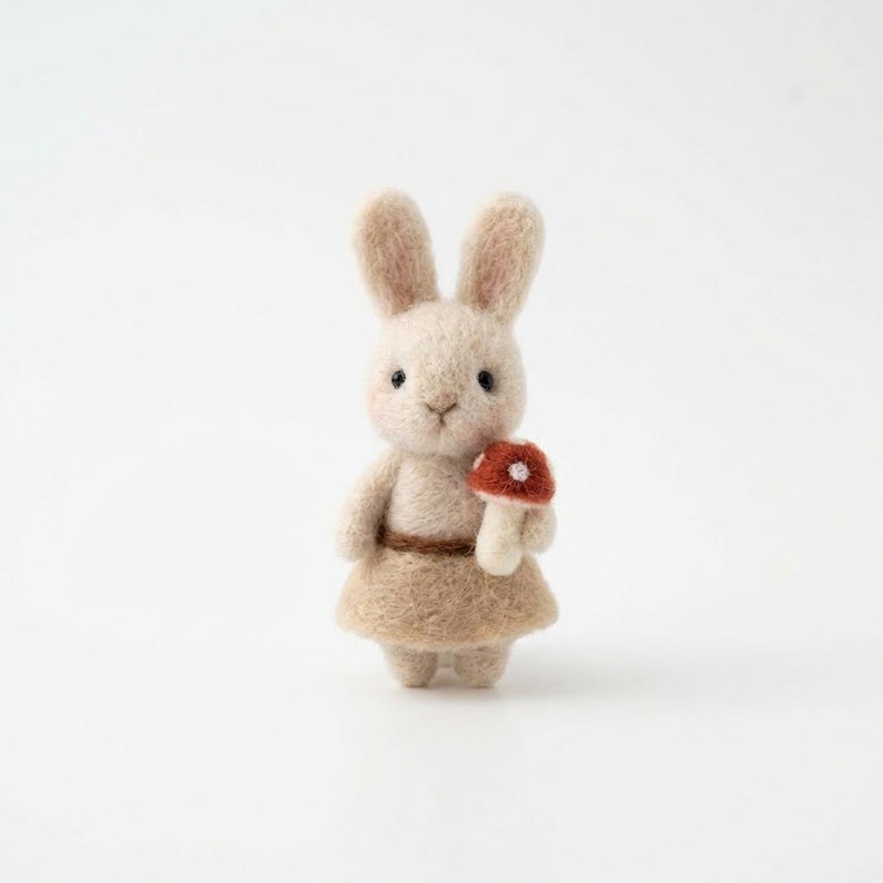 May include: A handmade felt bunny figurine in a light beige color, wearing a matching skirt and holding a small red and white mushroom. The bunny has long ears and a sweet expression, perfect for a nursery or as a whimsical gift.