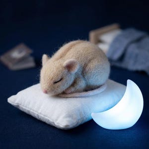 May include: A beige felt mouse figurine, crafted by hand, asleep on a small white pillow. A glowing, crescent moon-shaped night light is positioned close by. The background is a dark blue, creating a cosy, whimsical atmosphere.