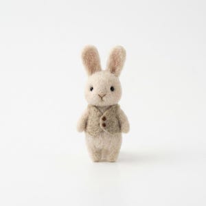 May include: A handcrafted, beige felt bunny figurine wearing a tan vest with two brown buttons. The bunny has upright ears, black eyes, and a small, stitched nose. The background is plain white.