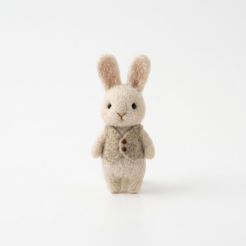 May include: A handcrafted, beige felt bunny figurine wearing a tan vest with two brown buttons. The bunny has upright ears, black eyes, and a small, stitched nose. The background is plain white.