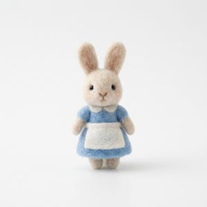 May include: A handcrafted felt bunny figurine. The bunny has beige fur, long ears, and black eyes. It wears a light blue dress with a white apron and collar. The bunny is standing upright against a white background.