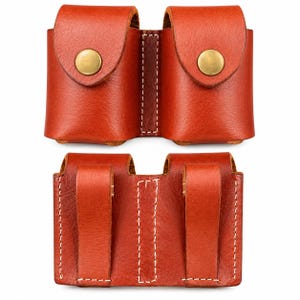 May include: A double pouch made of reddish-brown leather featuring two front flaps secured with brass-colored metal snap buttons. The back of the pouch displays two vertical leather belt loops stitched with white thread for attachment.