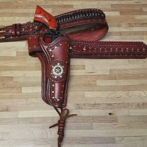 Op de afbeelding: Een handgemaakte, roodbruine leren holster- en riemset. De holster heeft een decoratief sterembleem en is versierd met klinknagels. De riem bevat lussen voor kogels en een gesp. De set is ontworpen voor een vuurwapen.