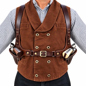 Könnte beinhalten: Eine braune zweireihige Weste mit Schalkragen und Messingknöpfen, getragen über einem hellblau-weiß gestreiften Hemd. Ein dunkelbraunes Schulterholster-System aus Leder ist über der Weste befestigt und hält zwei silberne Revolver an der Taille.