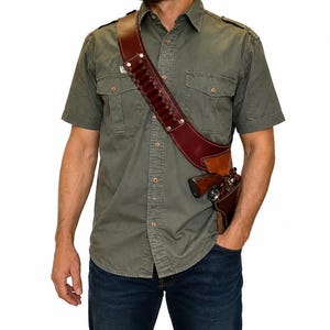 May include: A brown leather bandolier holster worn across the chest, featuring a series of loops for ammunition cartridges along the strap and a moulded holster pocket at the hip containing a metallic revolver with a wooden grip handle.
