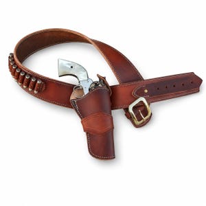 May include: A brown leather gun belt with a matching holster containing a silver revolver with a white pearlescent grip. The belt features a brass buckle and a series of loops holding metallic cartridges along the side of the leather strap.
