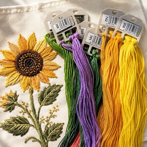 May include: A piece of off-white fabric featuring an embroidered sunflower with yellow petals and a dark brown centre, accompanied by green leaves and stems. Next to the embroidery are several skeins of embroidery floss in green, purple, and yellow.