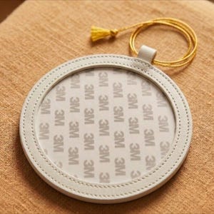 May include: A circular, off-white leather coaster with a stitched edge. It features a small loop and a gold tassel. The centre of the coaster has the repeated text "3M".
