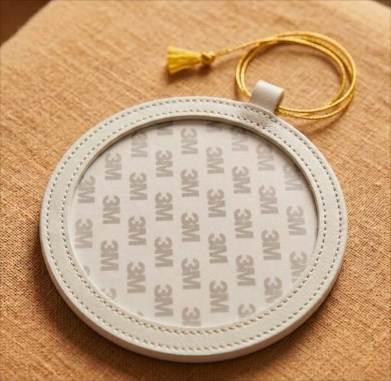 May include: A circular, off-white leather coaster with a stitched edge. It features a small loop and a gold tassel. The centre of the coaster has the repeated text "3M".