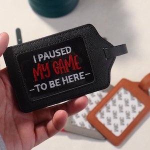 K&ouml;nnte beinhalten: Schwarzes Gep&auml;ckanh&auml;nger mit dem gestickten Text "I PAUSED MY GAME -TO BE HERE-". Das Etikett hat ein klares Fenster f&uuml;r eine ID-Karte und einen kleinen Griff. Weitere Gep&auml;ckanh&auml;nger sind im Hintergrund sichtbar.