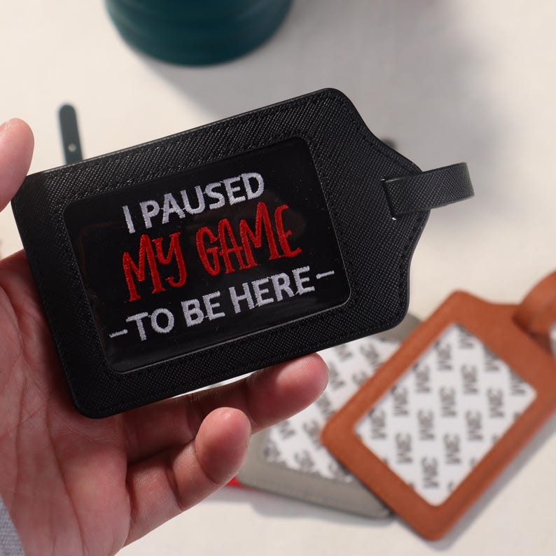 K&ouml;nnte beinhalten: Schwarzes Gep&auml;ckanh&auml;nger mit dem gestickten Text "I PAUSED MY GAME -TO BE HERE-". Das Etikett hat ein klares Fenster f&uuml;r eine ID-Karte und einen kleinen Griff. Weitere Gep&auml;ckanh&auml;nger sind im Hintergrund sichtbar.