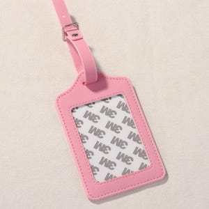 K&ouml;nnte beinhalten: Ein pinkfarbenes Gep&auml;ckanh&auml;nger mit einem transparenten Fenster und einem passenden Riemen. Der Anh&auml;nger hat eine rechteckige Form mit einem dekorativen oberen Rand und einer gen&auml;hten Bord&uuml;re. Der Riemen hat eine silberne Schnalle. Das Fenster zeigt den Text "3M".