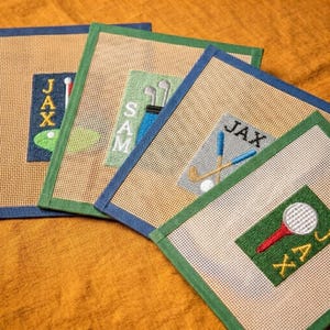 May include: Four square needlepoint canvases with tan mesh backgrounds and coloured borders. Each features embroidered golf-themed designs including golf clubs, balls, and the names JAX and SAM stitched in various coloured thread blocks.