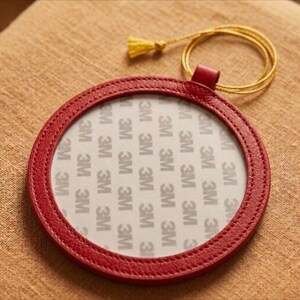 May include: A circular, red leather coaster with a stitched edge. The coaster features a clear centre with the text "3M" repeated. A gold-coloured cord with a tassel is attached to the coaster.
