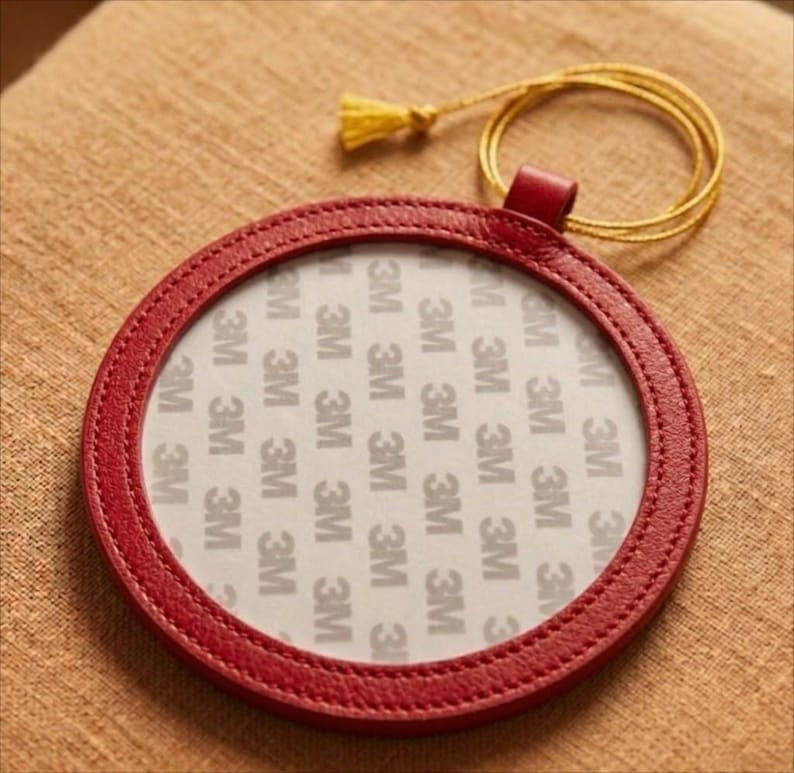 May include: A circular, red leather coaster with a stitched edge. The coaster features a clear centre with the text "3M" repeated. A gold-coloured cord with a tassel is attached to the coaster.