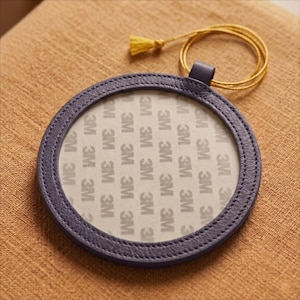 May include: A circular, navy blue leather frame with a gold tassel and cord. The frame features a stitched edge and a white interior with the text "3M" repeated. It rests on a brown surface.