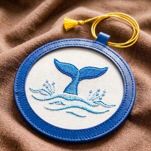 May include: A circular embroidered design featuring a blue whale tail emerging from waves. The design is set in a blue frame with a yellow cord and tassel attached. The design is on a white background.