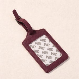 K&ouml;nnte beinhalten: Ein weinrotes Gep&auml;ckanh&auml;nger mit strukturierter Oberfl&auml;che und einem transparenten Fenster. Der Anh&auml;nger hat einen Riemen mit Schnalle und der Text "3M" ist im Fenster sichtbar.