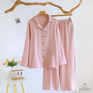 Może przedstawiać: Jasnor&oacute;żowy zestaw piżamy, składający się z koszuli zapinanej na guziki z długim rękawem i pasujących spodni, wisi na drewnianym wieszaku. Koszula posiada mały haftowany wz&oacute;r misia i kontrastowe ciemnobrązowe wykończenie wok&oacute;ł kołnierza. Tkanina ma teksturowany wygląd.