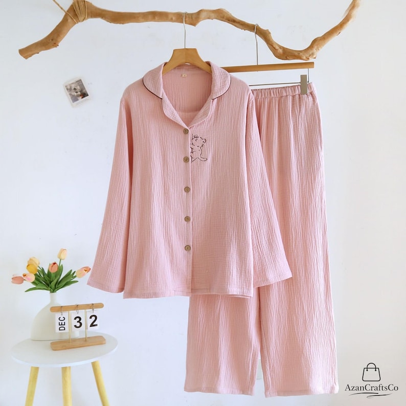 Może przedstawiać: Jasnor&oacute;żowy zestaw piżamy, składający się z koszuli zapinanej na guziki z długim rękawem i pasujących spodni, wisi na drewnianym wieszaku. Koszula posiada mały haftowany wz&oacute;r misia i kontrastowe ciemnobrązowe wykończenie wok&oacute;ł kołnierza. Tkanina ma teksturowany wygląd.