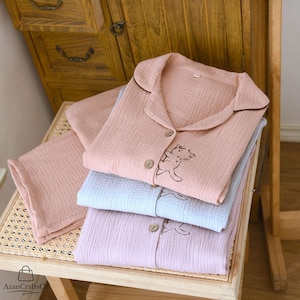 Może przedstawiać: Ułożone w stos zestawy piżam w pastelowych kolorach. Każdy zestaw zawiera koszulę zapinaną na guziki z haftem przedstawiającym misia z kresk&oacute;wki i pasujące spodnie. Piżamy są w odcieniach r&oacute;żu, błękitu i lawendy, z brązowymi wykończeniami i guzikami. Wykonane z miękkiej, teksturowanej tkaniny.