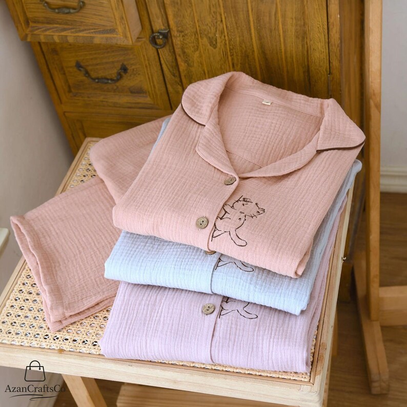 Może przedstawiać: Ułożone w stos zestawy piżam w pastelowych kolorach. Każdy zestaw zawiera koszulę zapinaną na guziki z haftem przedstawiającym misia z kresk&oacute;wki i pasujące spodnie. Piżamy są w odcieniach r&oacute;żu, błękitu i lawendy, z brązowymi wykończeniami i guzikami. Wykonane z miękkiej, teksturowanej tkaniny.