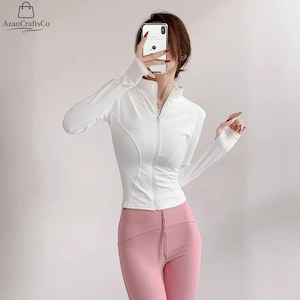 Könnte beinhalten: Weiße Sportjacke mit Reißverschluss, langen Ärmeln und Daumenlöchern. Die Jacke hat ein figurbetontes Design mit geschwungenen Einsätzen. Kombiniert mit rosa Leggings. Der Markenname "AzanCraftsCo" ist oben links sichtbar.
