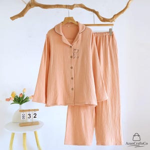 Może przedstawiać: Brzoskwiniowy zestaw piżamowy z koszulą zapinaną na guziki i pasującymi spodniami. Koszula ma mały haftowany wz&oacute;r i ciemnobrązowe wykończenie na kołnierzu. Zestaw wisi na drewnianym wieszaku na białym tle.