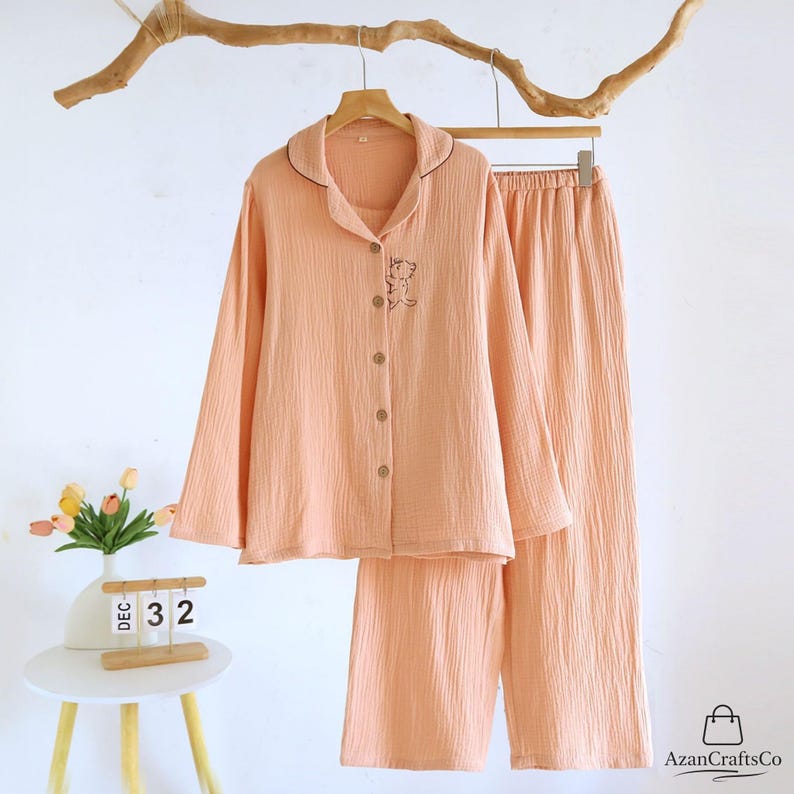 Może przedstawiać: Brzoskwiniowy zestaw piżamowy z koszulą zapinaną na guziki i pasującymi spodniami. Koszula ma mały haftowany wz&oacute;r i ciemnobrązowe wykończenie na kołnierzu. Zestaw wisi na drewnianym wieszaku na białym tle.