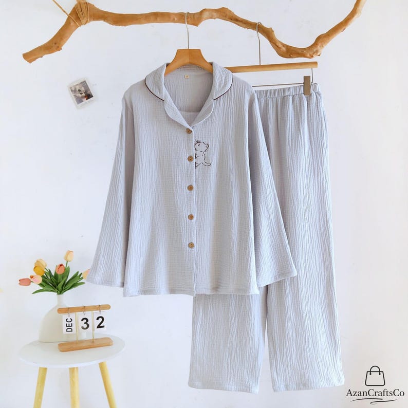 Może przedstawiać: Jasnoszary zestaw piżamowy z koszulą zapinaną na guziki z długim rękawem i pasującymi spodniami. Koszula ma mały haftowany wz&oacute;r misia i brązowe guziki. Zestaw wisi na drewnianym wieszaku.