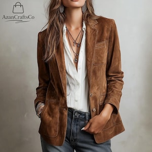 Puede incluir: Una blazer de ante marrón con dos bolsillos delanteros y cierres de botón. La blazer se lleva sobre una camisa blanca abotonada y vaqueros azules. La imagen también muestra collares superpuestos y pendientes.