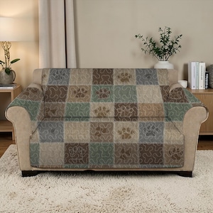 May include: A beige sofa is covered with a patchwork style throw. The throw features squares of various colours, including green, brown, and beige, with paw print patterns. The sofa sits on a fluffy, white rug.